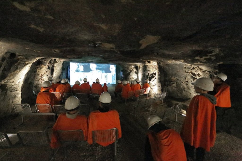 Mehrere Besucher schauen unter Tage auf einer großen Leinwand einen Film.