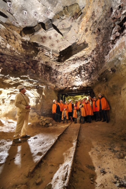 Die Besucher stehen im Bergwerk. Im Vordergrund sind Gleise zu sehen. Ein Führer in weiß steht vorne links im Bild.