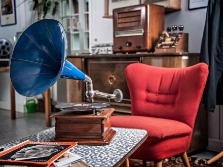Ein roter Sessel aus den 60ern steht im Raum. Im Vordergrund steht ein Grammophon mit blauem Horn auf dem Tisch, davor eine Ausgabe des Spiegels mit einem schwarz-weißen Portrait darauf. Im Hintergrund findet sich ein altes Radio im Holzgehäuse sowie eine Rechenmaschine oder Kasse.